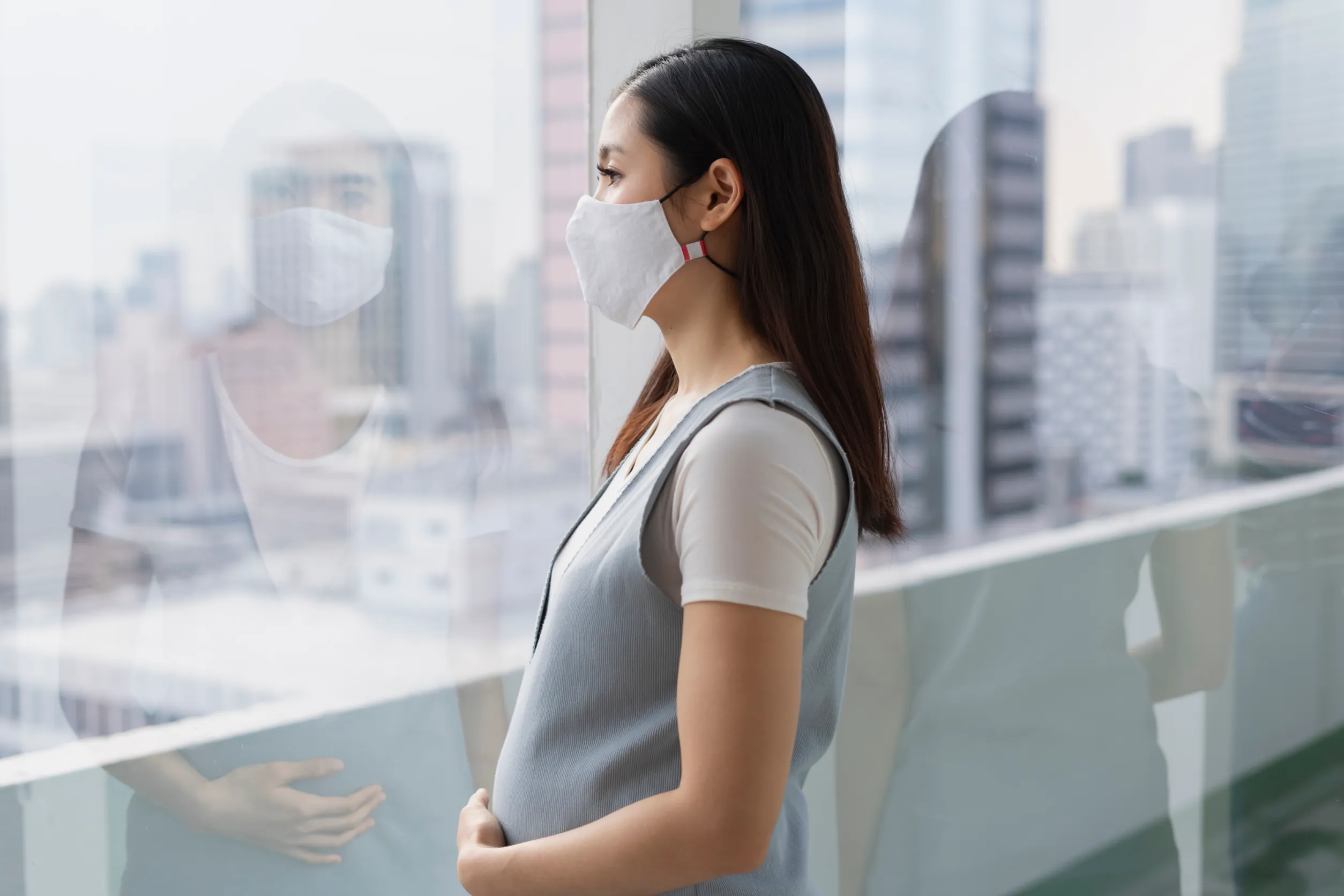 Beautiful pregnant asian woman wearing mask looking outside the window. Covid-19 outbreak.